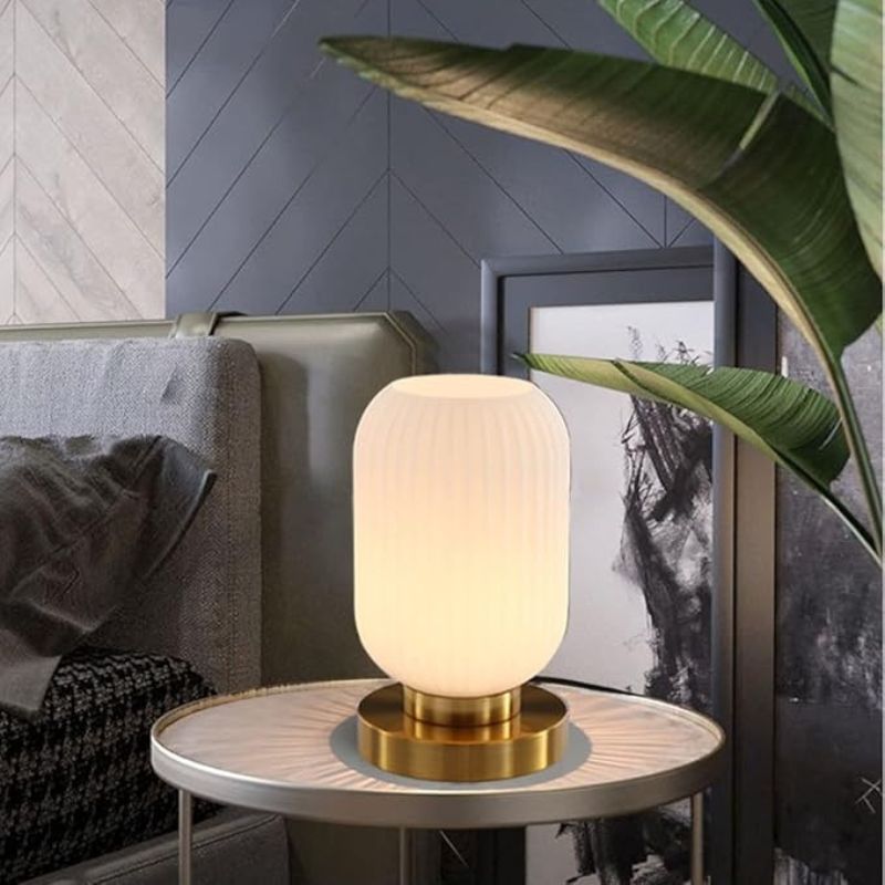 A lit, ribbed white glass table lamp with a gold base sits on a small round table next to a bed and framed artwork. Part of a large green plant is visible in the foreground. This could be an Auwieou Mid Century Modern Table Lamp.