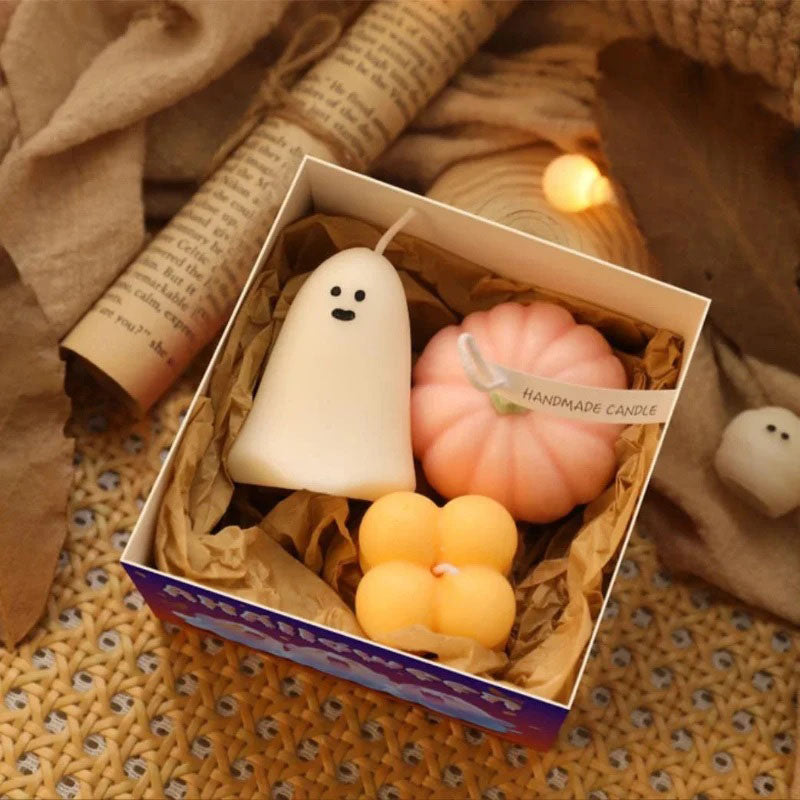 A gift box contains three Halloween-themed candles nestled in brown crinkle-cut paper. Inside are a white ghost-shaped candle with two black dot eyes, a pink pumpkin-shaped candle with a white string tied around it and a "Handmade Candle" tag, and two stacked orange cube-shaped candles. The box sits on a textured surface with soft, warm-toned string lights scattered around it. A rolled-up piece of paper with printed text is visible in the upper left corner.