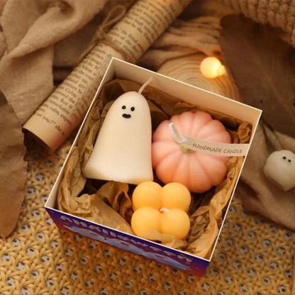A gift box contains three Halloween-themed candles nestled in brown crinkle-cut paper. Inside are a white ghost-shaped candle with two black dot eyes, a pink pumpkin-shaped candle with a white string tied around it and a "Handmade Candle" tag, and two stacked orange cube-shaped candles. The box sits on a textured surface with soft, warm-toned string lights scattered around it. A rolled-up piece of paper with printed text is visible in the upper left corner.