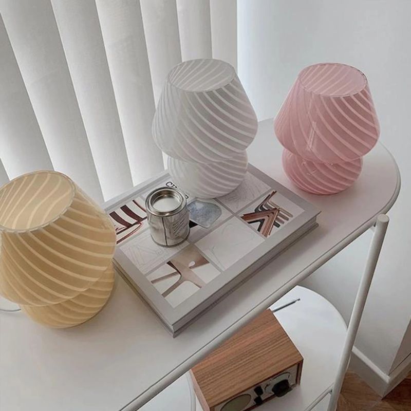 Three striped glass mushroom lamps in cream, white, and pink sit atop a modern white console table. A stylish book and a small silver tin add to the minimalist decor, while natural light filters through vertical blinds in the background. The soft pastel tones and artistic design create a serene, contemporary aesthetic.