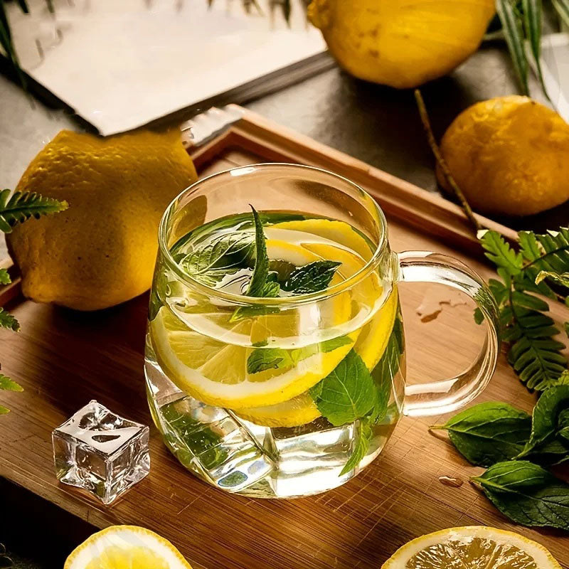 A glass mug of lemon and mint water with an ice cube sits on a wooden board surrounded by whole and sliced lemons.