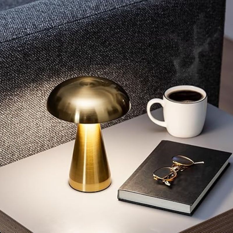 A gold, mushroom-shaped lamp with a warm glow sits on a light-colored surface next to a dark-covered book, a pair of gold-framed glasses, and a white mug of coffee, with a portion of a textured gray couch visible in the background, creating a cozy and inviting scene.