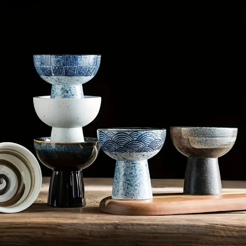 Aesthetic pedestal bowls in various ceramic designs, featuring speckled, glazed, and traditional blue patterns. Stacked and arranged on a wooden surface for an elegant display.