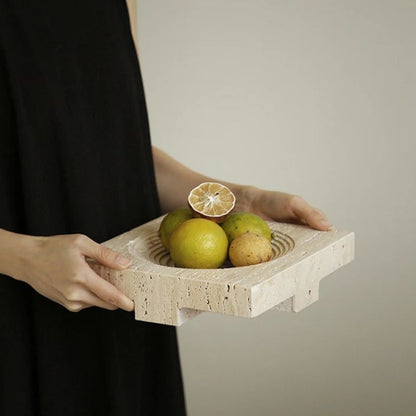 A person in a black dress holds a textured Travertine Stone Tray adorned with lemons and potatoes. A halved lime elegantly sits atop the ensemble, creating an organic texture that enhances the visually appealing arrangement against a plain backdrop, perfect for home styling.