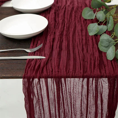 Burgundy crinkled table runner on a rustic wooden table, elegantly set with white plates, silverware, and greenery accents.