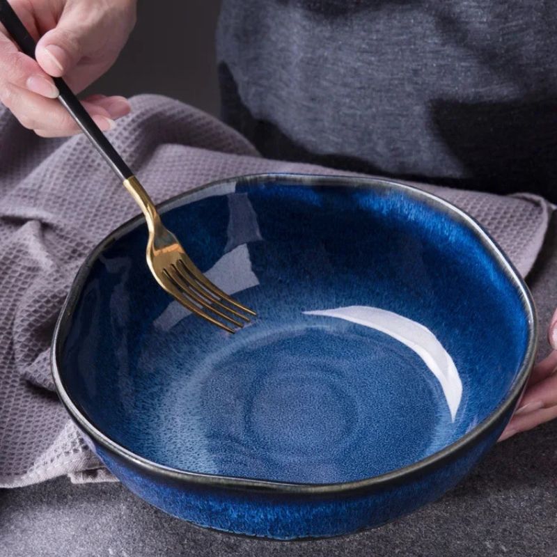 Close-up of a person holding a gold and black fork above a deep blue ceramic wave bowl with a glossy finish. The bowl's organic shape and rich glaze contrast against a textured gray fabric background.