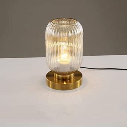 A lit, ribbed amber glass table lamp with a gold base sits on a white surface against a plain gray background. This could be a TaleTre Modern Glass Table Lamp.