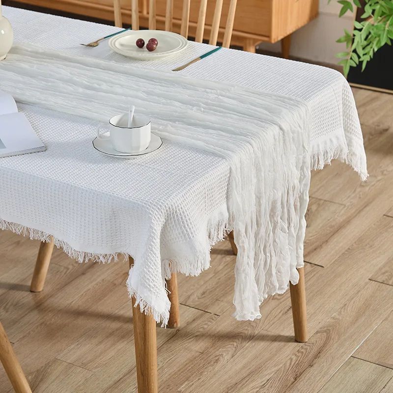 White crinkled table runner over a textured tablecloth on a wooden dining table, set with a teacup, plate, and cutlery in a cozy setting.