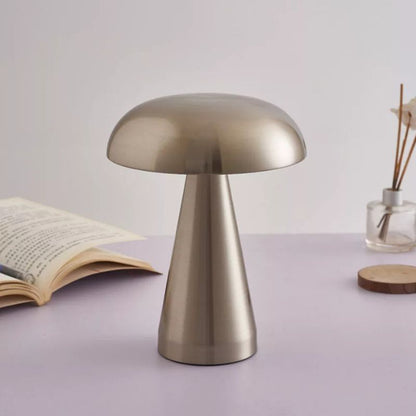 A silver, mushroom-shaped lamp stands prominently on a light-colored surface, with an open book to its left and a small glass vase with sticks and a wooden disc to its right, all set against a soft, light background.
