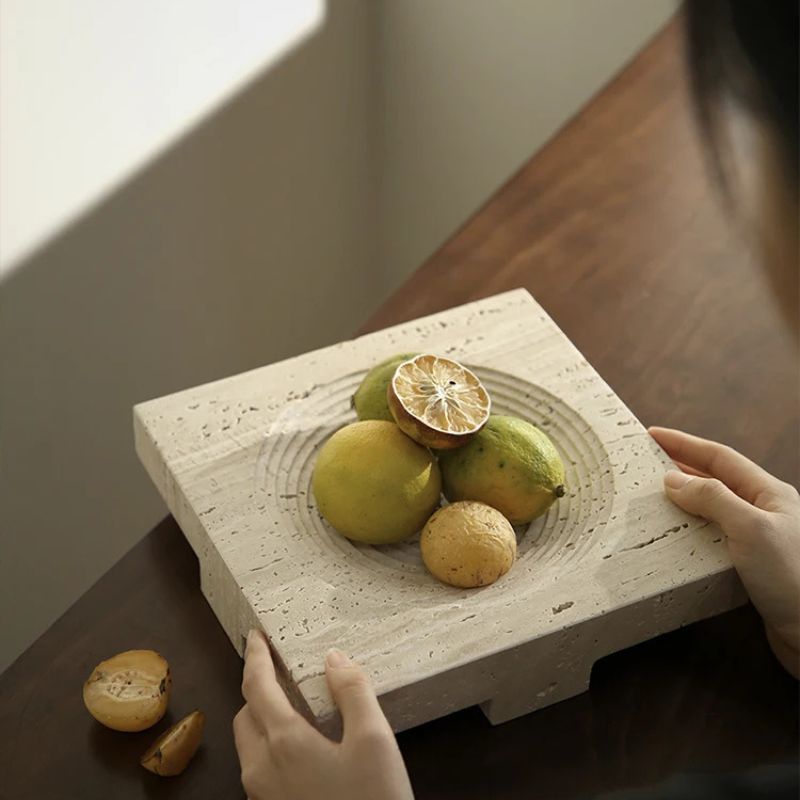 A person holds a travertine stone slab with a circular indentation, displaying limes and a sliced lime on top. A couple of lemons rest on the dark wooden table nearby, enhancing the natural beauty. Soft natural light creates a warm atmosphere perfect for elegant home styling.