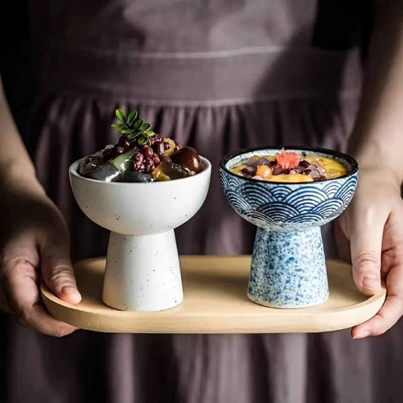 A person holds a wooden tray adorned with two ceramic dessert bowls, perfect for sweet moments. The left is white with assorted toppings, while the right showcases a blue and white wave pattern, cradling a fruity dessert topped with red beans—truly dessert time bliss.