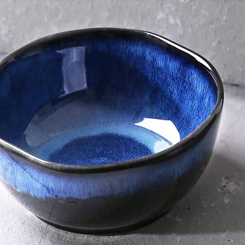 Handmade ceramic bowl with a deep blue gradient glaze on the interior and a glossy black exterior, set against a textured concrete background.