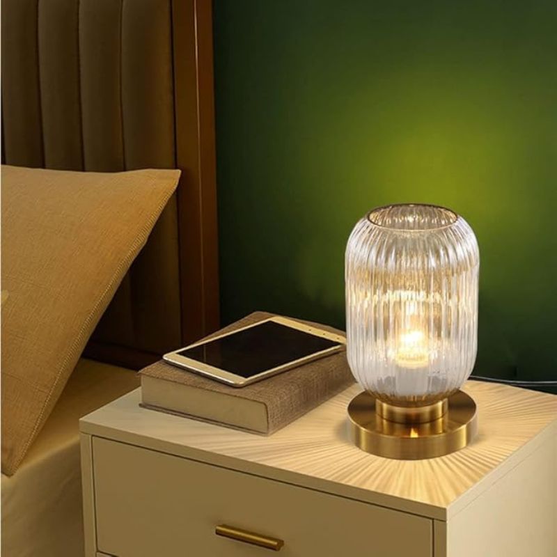 A lit ribbed glass table lamp with a gold base sits on a white nightstand next to a book and a tablet. A bed with a brown headboard and pillow is partially visible to the left, and a dark green wall is in the background. This could be an Auwieou Mid Century Modern Table Lamp.