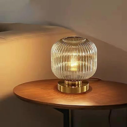 A lit, ribbed amber glass mini table lamp with a gold base sits on a round wooden table. A blurred light-colored sofa is partially visible in the background.
