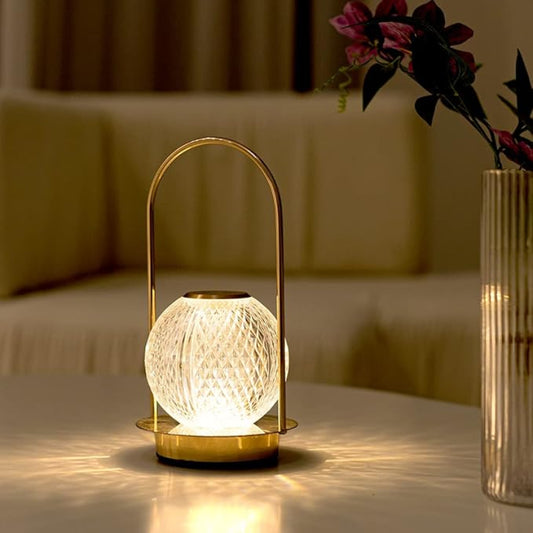 A lit modern lamp with a crystal-like spherical shade and a gold-colored metal base and handle, sitting on a white table. A vase of flowers and a sofa are in the soft-focus background.