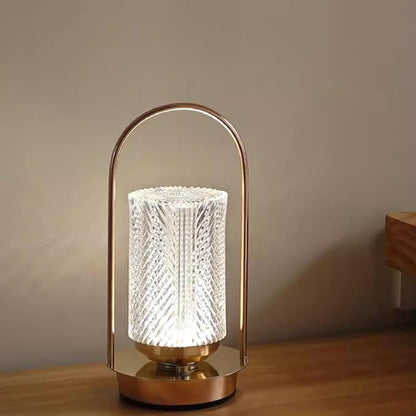 A lit modern lantern-style lamp with a textured cylindrical glass shade and a gold-toned base and handle, sitting on a wooden surface.