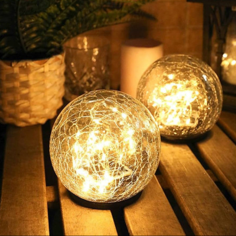 Two solar-powered crackle glass globe lights emit a warm white glow while placed on a wooden surface. The ambient lighting enhances the cozy and inviting atmosphere, with decorative elements like a woven basket and glassware in the softly lit background.