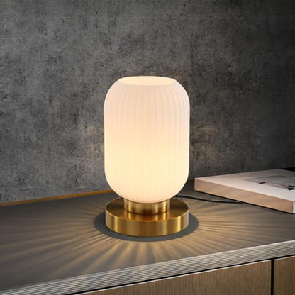A lit, ribbed white glass table lamp with a gold base sits on a dark gray surface, casting light patterns. A stack of papers rests behind it against a textured gray wall.