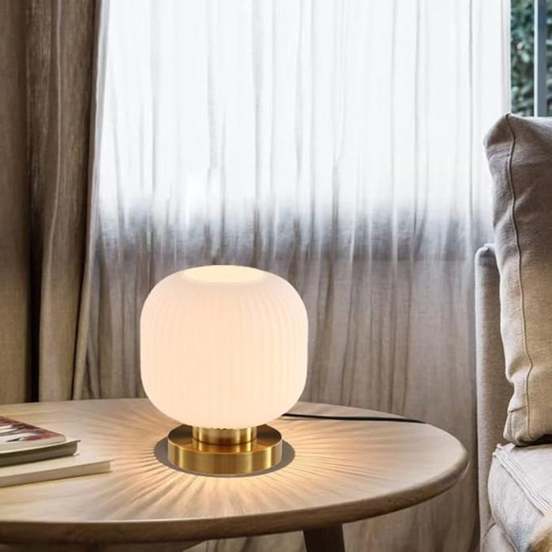 Lit white glass lamp with a gold base on a round table near books and a sofa.