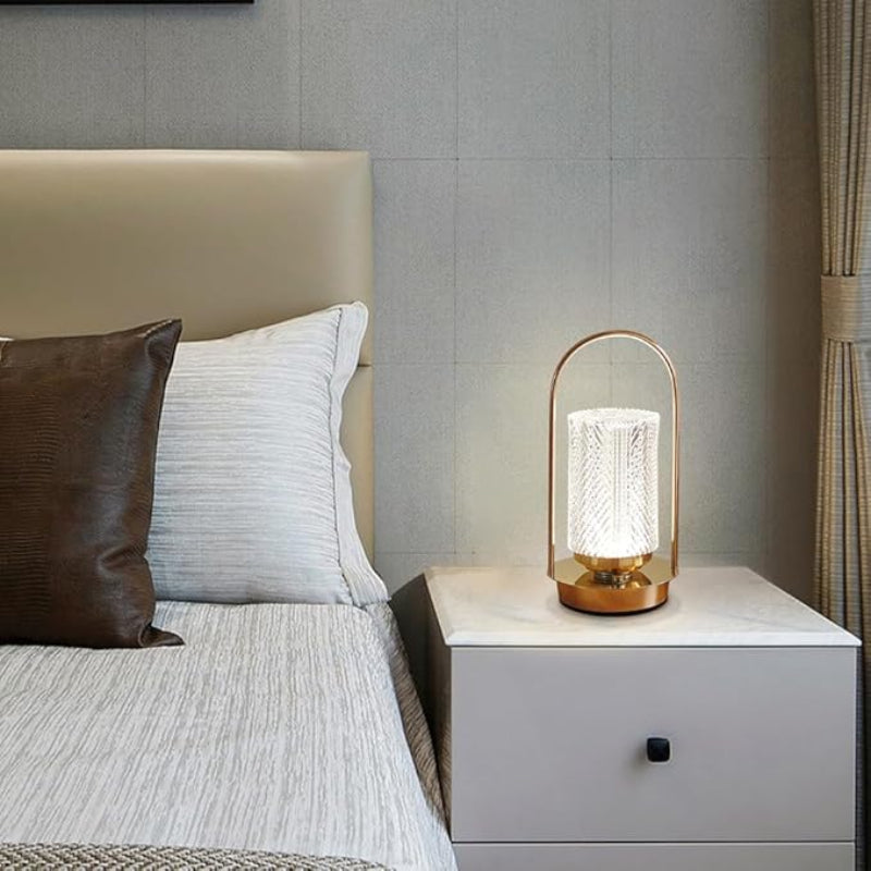 A lit modern lantern-style lamp with a textured cylindrical shade and a gold-toned base, sitting on a white marble nightstand next to a bed.