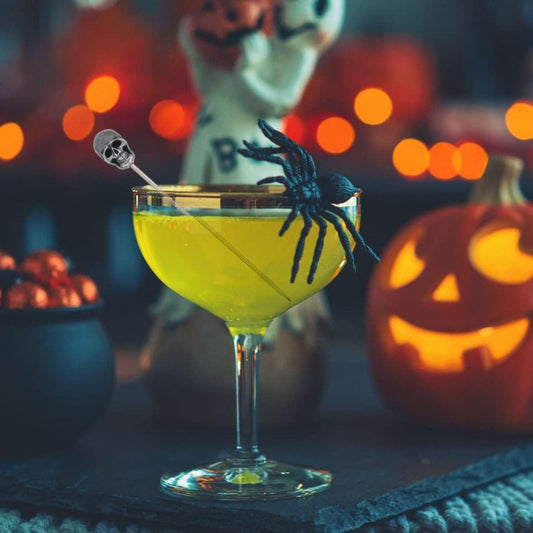 A yellow Halloween cocktail in a stemmed glass with a skull pick, spider decoration, and jack-o'-lantern background.