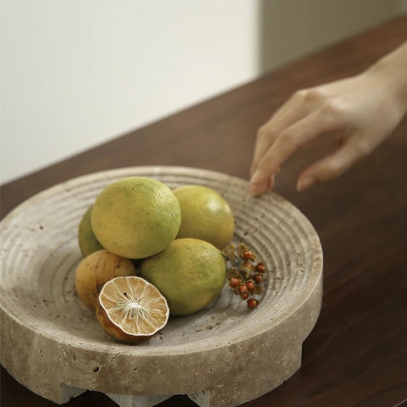 A hand reaches towards a round travertine stone plate with several lemons and a halved lemon. Small orange fruits are beside them on the wooden surface. The scene is softly lit, highlighting the natural beauty in this warm and stylish home setting.