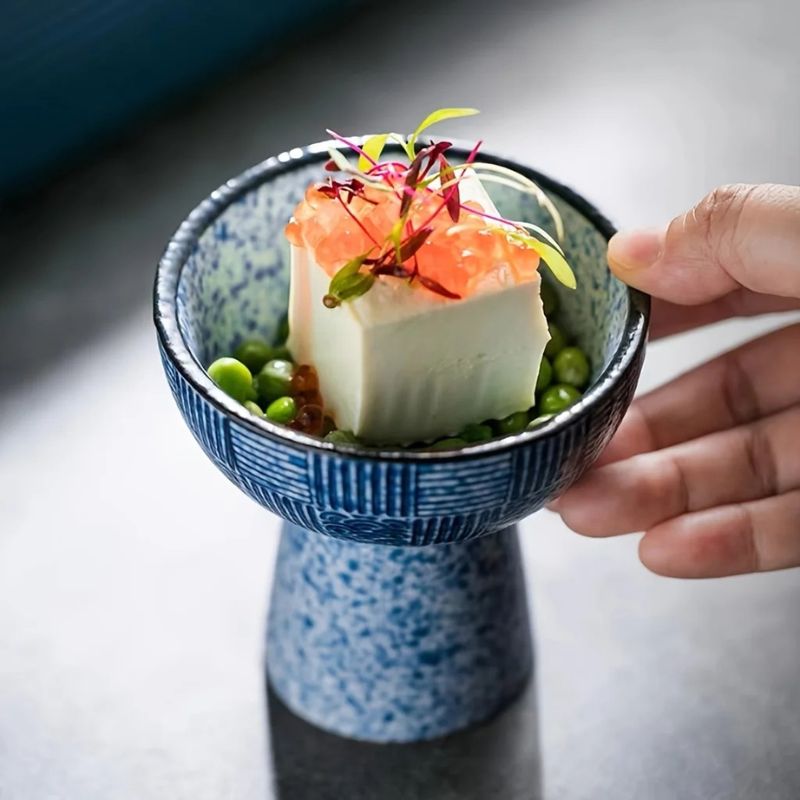 A hand holding a versatile ceramic bowl brimming with tofu garnished with orange roe and microgreens, all nestled on a bed of green peas. Perfect for any course, even dessert time.