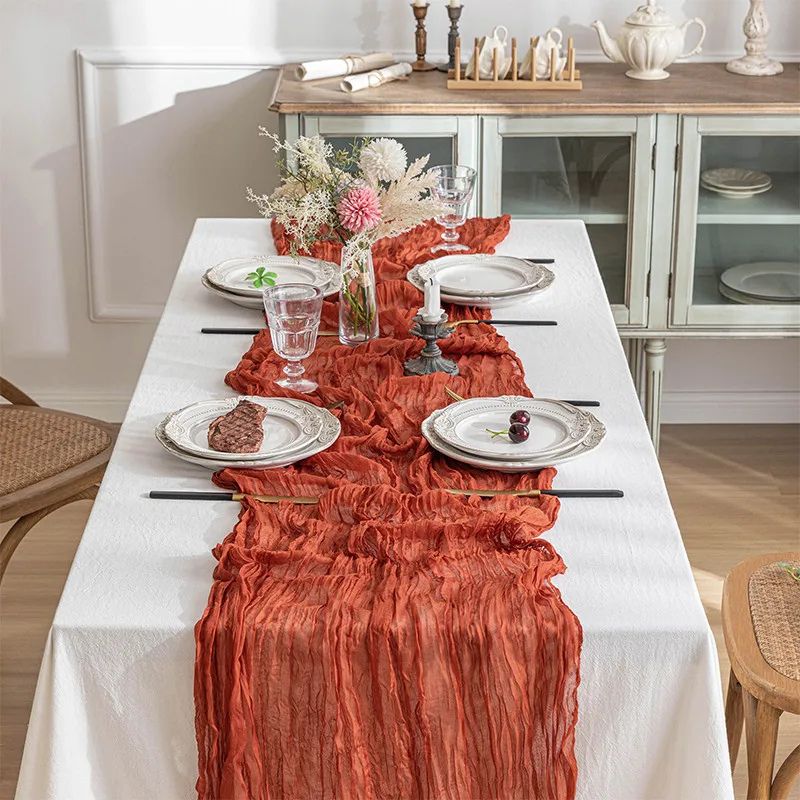 Rust-colored crinkled table runner on a white table, set with vintage plates, black chopsticks, glassware, and a floral centerpiece.