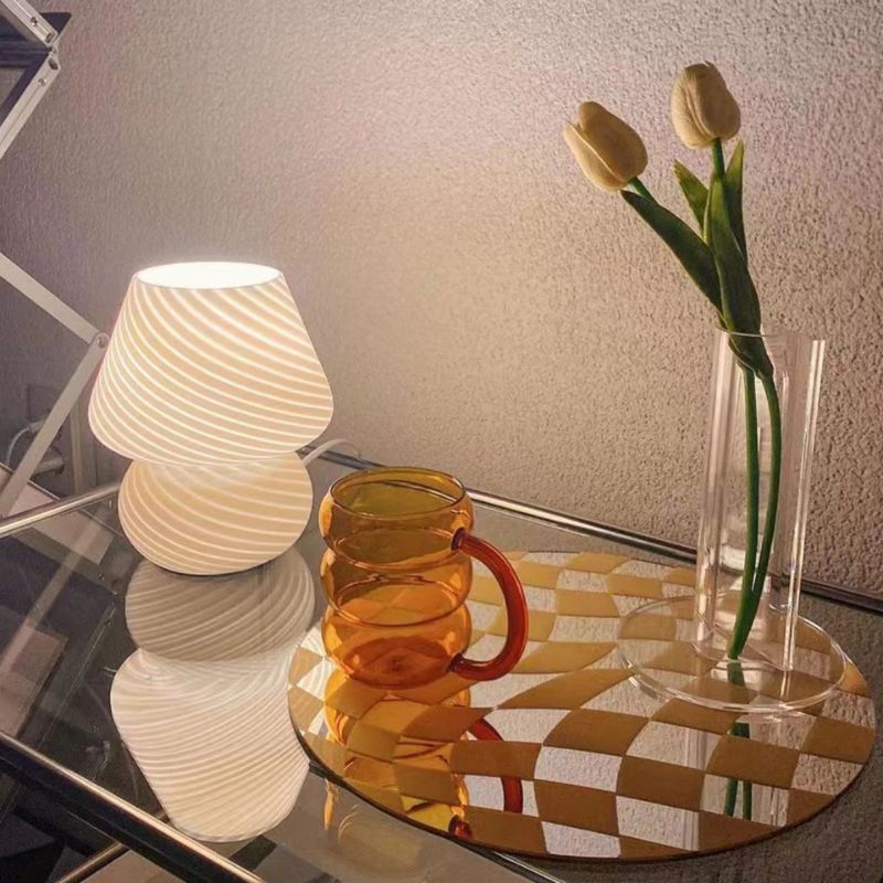 A modern dimmable mushroom lamp with a striped, translucent Murano-style shade emits a warm glow on a glass bedside table. Beside it, an amber-toned glass mug and a clear vase with two white tulips create a cozy and stylish aesthetic. The scene reflects softly on the tabletop, enhancing the elegant ambiance.