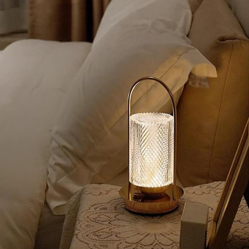 A lit bedside lamp with a textured cylindrical shade and a gold base, placed on a nightstand next to a bed.