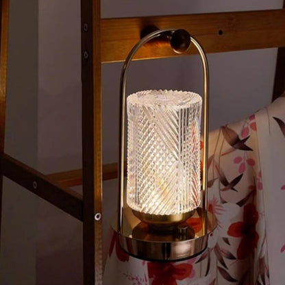 A lit modern lamp with a textured cylindrical glass shade and a gold-toned base and handle, hanging from a wooden shelf.