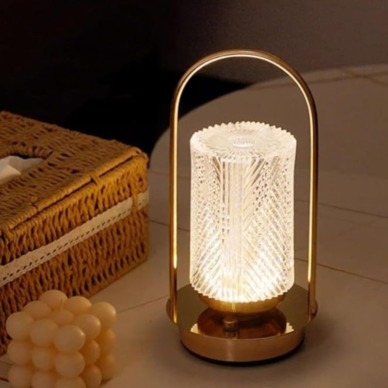 A lit modern lantern-style lamp with a textured glass shade and a gold-toned base, sitting on a surface next to a woven box and a bubble candle.
