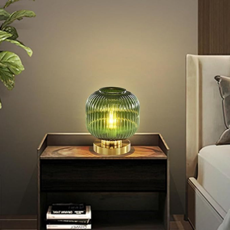 A lit DoungRos green glass table lamp with a gold base sits on a dark wood nightstand next to a bed. A stack of books is on the lower shelf of the nightstand, and a leafy plant is partially visible in the upper left corner. The wall behind the nightstand is a muted beige color.