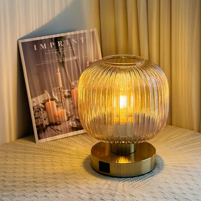 A lit, ribbed amber glass mini table lamp with a gold base sits on a textured surface next to a magazine titled "IMPRINT". The background features a softly lit, draped curtain.