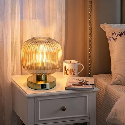 A lit, ribbed amber glass mini table lamp with a gold base sits on a white nightstand next to a white mug with the word "love" written on it and a small book. The nightstand is next to a bed with neutral-toned bedding and decorative pillows. Sheer curtains hang in the background.