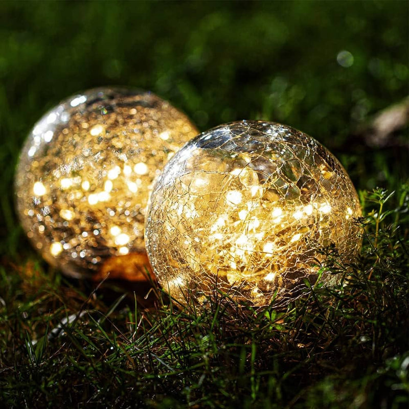 Two solar-powered crackle glass globe lights glowing warmly on a grassy surface at night. The LED lights inside the glass spheres create a beautiful, decorative ambiance, enhancing the outdoor setting with a cozy and magical atmosphere.