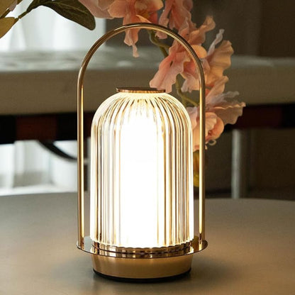 Gold table lamp with a soft glow on a surface, with flowers in the background.