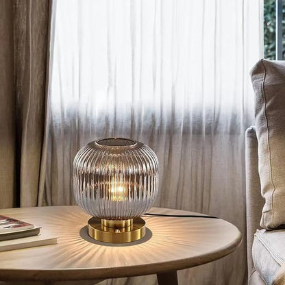 A lit, ribbed smoky gray glass mini table lamp with a gold base sits on a round wooden table next to a stack of books and a beige armchair. A window with sheer curtains is in the background.