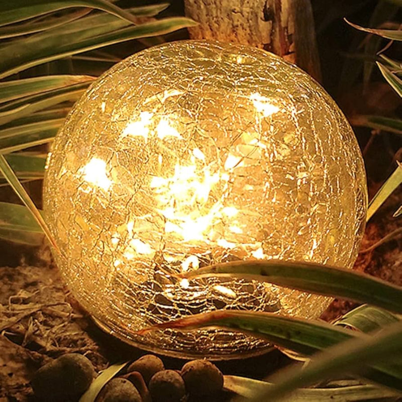 A glowing solar-powered crackle glass globe light placed among green leaves and small rocks in an outdoor garden setting at night. The warm LED lights inside the glass sphere create a cozy and decorative ambiance.