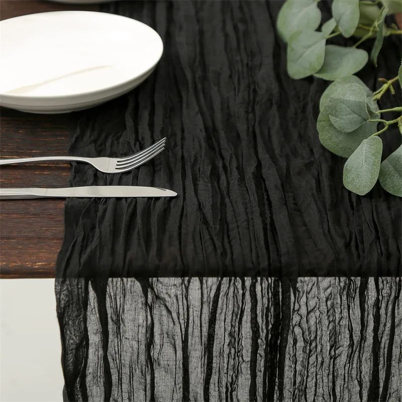 Black crinkled table runner draped over a wooden table, set with white plates, silver cutlery, and green foliage.