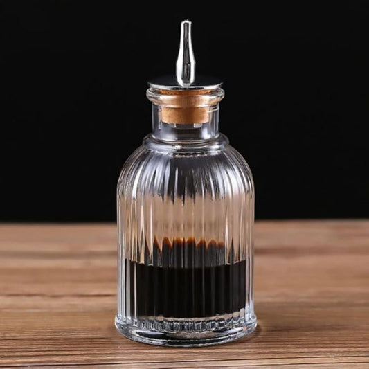 A ribbed glass bitters bottle with a metal pour spout and cork stopper, partially filled with dark liquid, sits on a wooden surface against a black background.