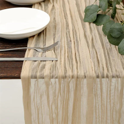 Beige crinkled table runner draped over a wooden table, set with white plates, silver cutlery, and green foliage.