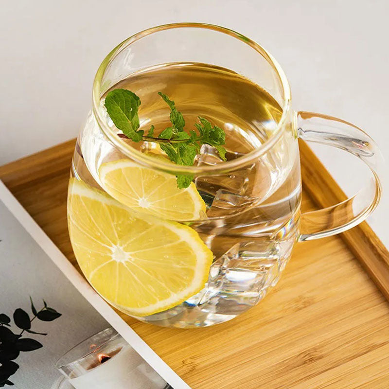 A close-up of a clear glass mug containing ice cubes, lemon slices, and mint leaves on a bamboo tray.