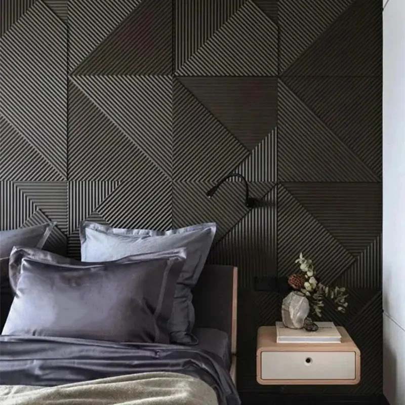 A modern bedroom features a bed with gray bedding and a light wood nightstand next to it, against a dark, geometric wood-paneled wall.