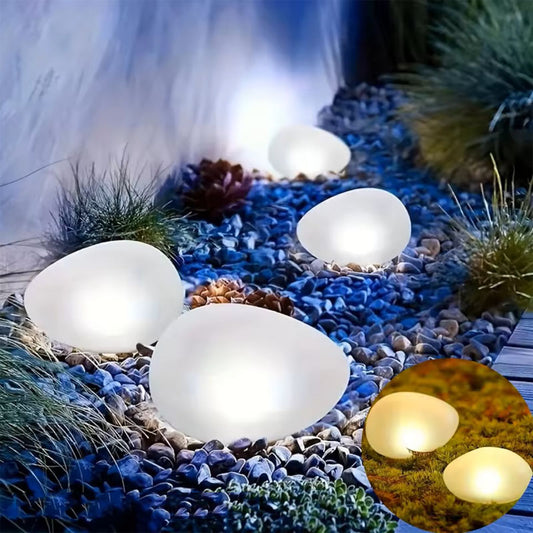 Several illuminated, smooth, white stone-shaped lights nestled in a rocky garden bed. An inset image shows two of the lights from a different angle on a mossy surface.