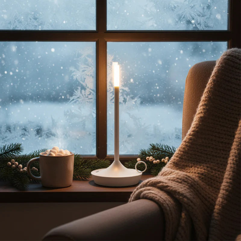 Cozy winter scene with a lit candle, mug of hot chocolate, and blanket by a snowy window.