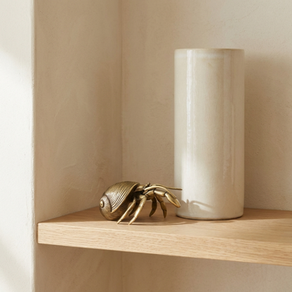 Gold crab figurine and white cylindrical vase on a wooden shelf against a beige wall.