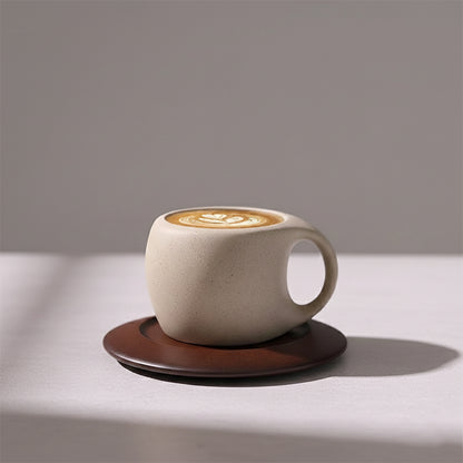 Rustic beige ceramic espresso cup with a wide handle, sitting on a dark brown saucer, containing coffee with latte art.