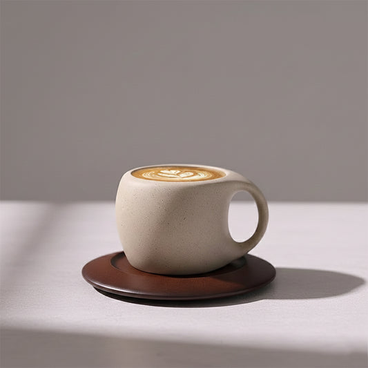 Rustic beige ceramic espresso cup with a wide handle, sitting on a dark brown saucer, containing coffee with latte art.