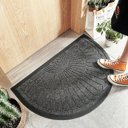 Half-circle grey welcome mat with a feather-like pattern in front of a wooden door, with a person's feet wearing orange sneakers visible.